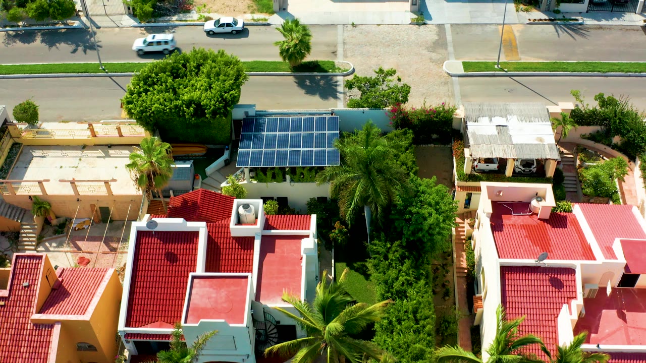 Aerial frame over a San Jose del Cabo residential block showing red tiled roofs, palms, a solar panel array, a paved street, and a white car near the upper road with no visible people.