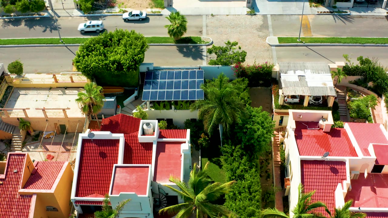 Top-down San Jose del Cabo neighborhood view with the white car farther along the upper street, red and pink rooftops below, solar panels at center right, and no visible people.