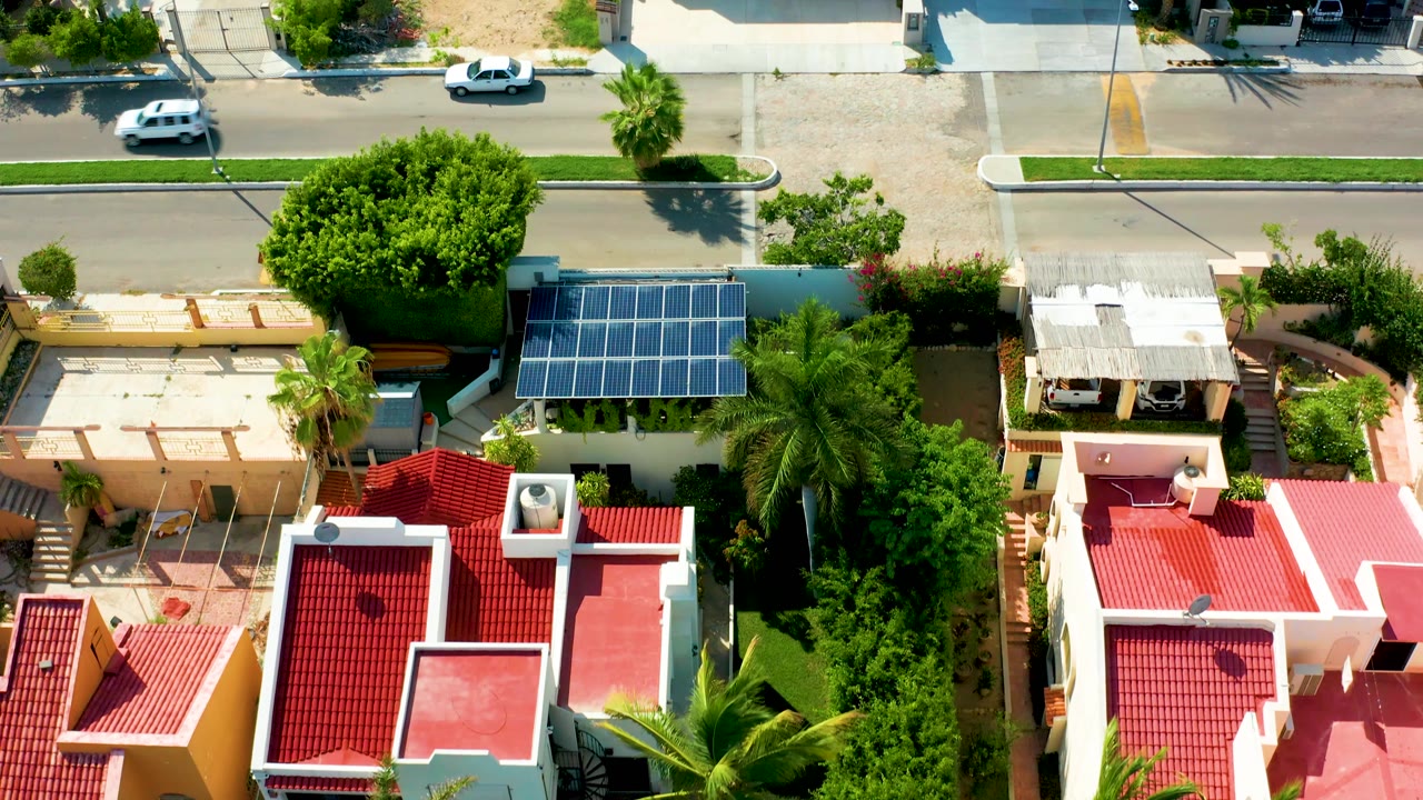 Aerial San Jose del Cabo frame showing a white car near the top road, a large green tree, blue solar panels, red rooftops, palms, and no visible people.