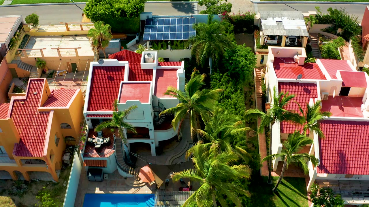 Overhead frame in San Jose Del Cabo showing yellow and pink homes with red roofs, tall palms, dense trees, and rooftop solar panels near the upper street, with no visible people.