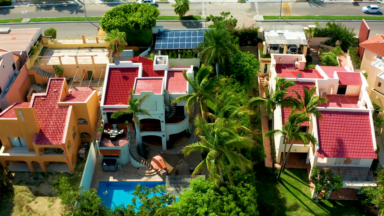 Aerial frame in San Jose Del Cabo showing the red-roofed homes slightly closer, with palms filling the center and the blue swimming pool low in frame, no visible people.