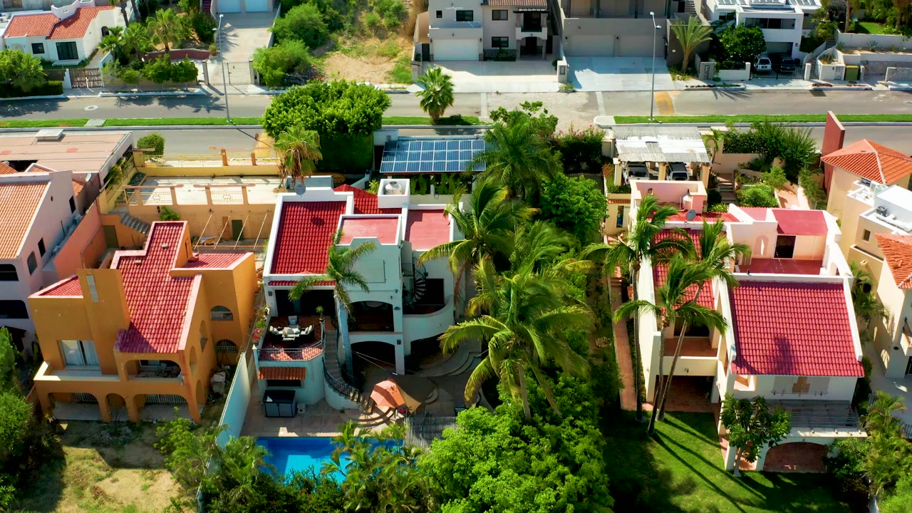 A drone aerial over San Jose del Cabo homes shows red tile roofs, tall palms, a blue backyard pool at lower left, a street at the top, and no visible people.