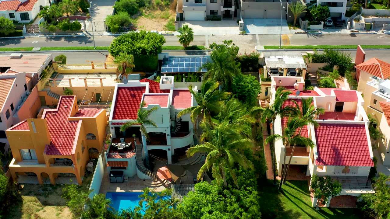 Aerial of a residential street, solar panels, palms, roofs, and pool.