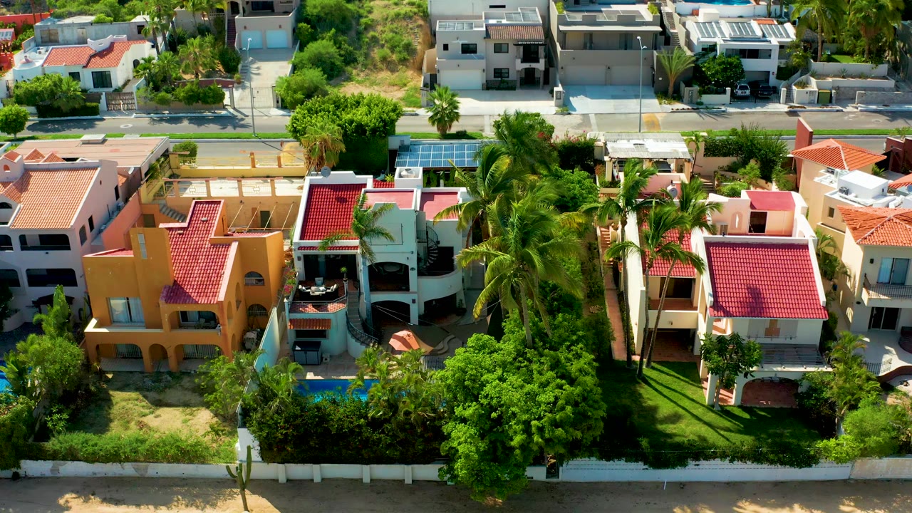Overhead drone frame in San Jose Del Cabo showing red-roofed houses, palms, blue pools, lawns, trees, a paved road, parked cars, and no visible people.