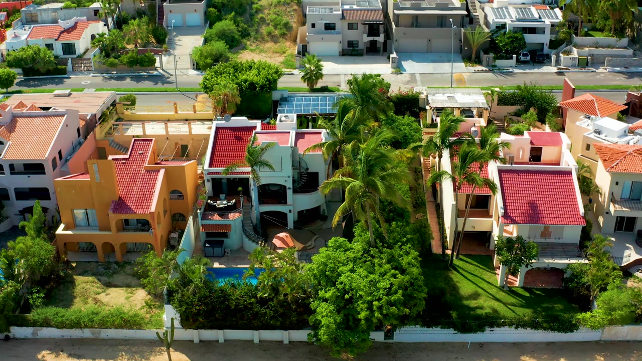 Top-down San Jose Del Cabo frame showing a cluster of homes with red tile roofs, tall palms, large green trees, turquoise pools, road edges, and no visible people.