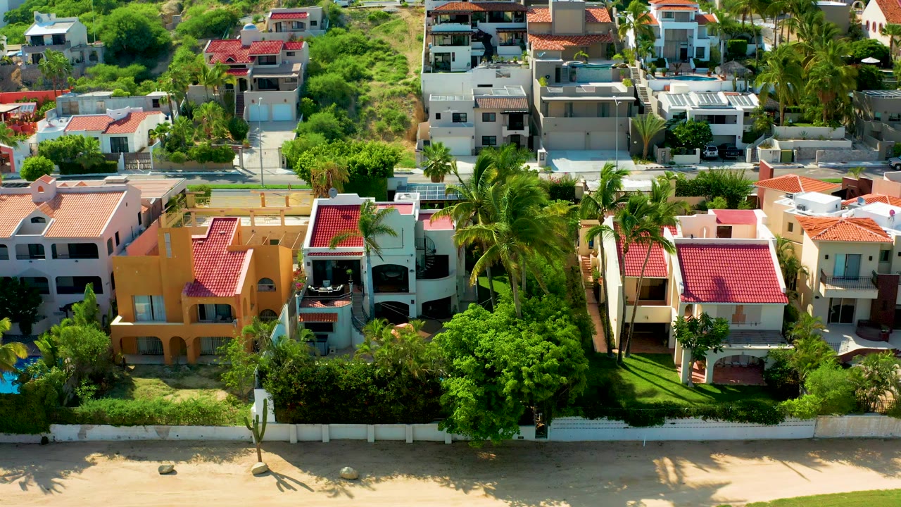 Aerial frame from San Jose Del Cabo showing sandy beach, a white wall, dense green trees, palms, an orange beachfront house, red-tile roofs, and no visible people.