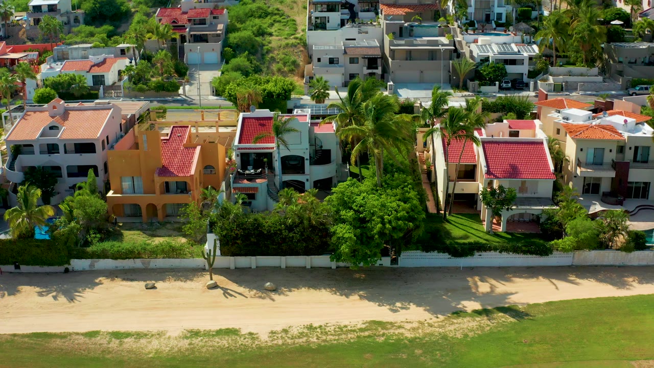 San Jose Del Cabo coastal residences fill the upper frame while palm trees, boundary walls, pale beach sand, shallow water, and no visible people occupy the shore below.
