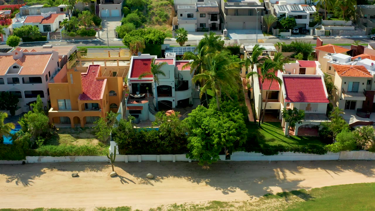 Aerial frame from San Jose Del Cabo showing a sandy beach and grass foreground below walled beachfront homes with palms and red roofs, with no visible people.
