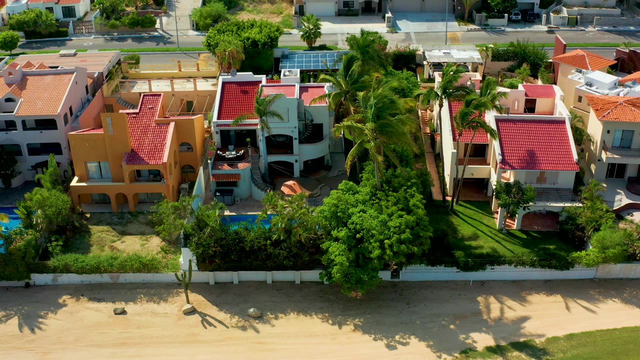 Aerial frame in San Jose Del Cabo, Baja California Sur, showing red-roof beachfront residences, blue pools, palm trees, a white fence, sandy shore, and no visible people.