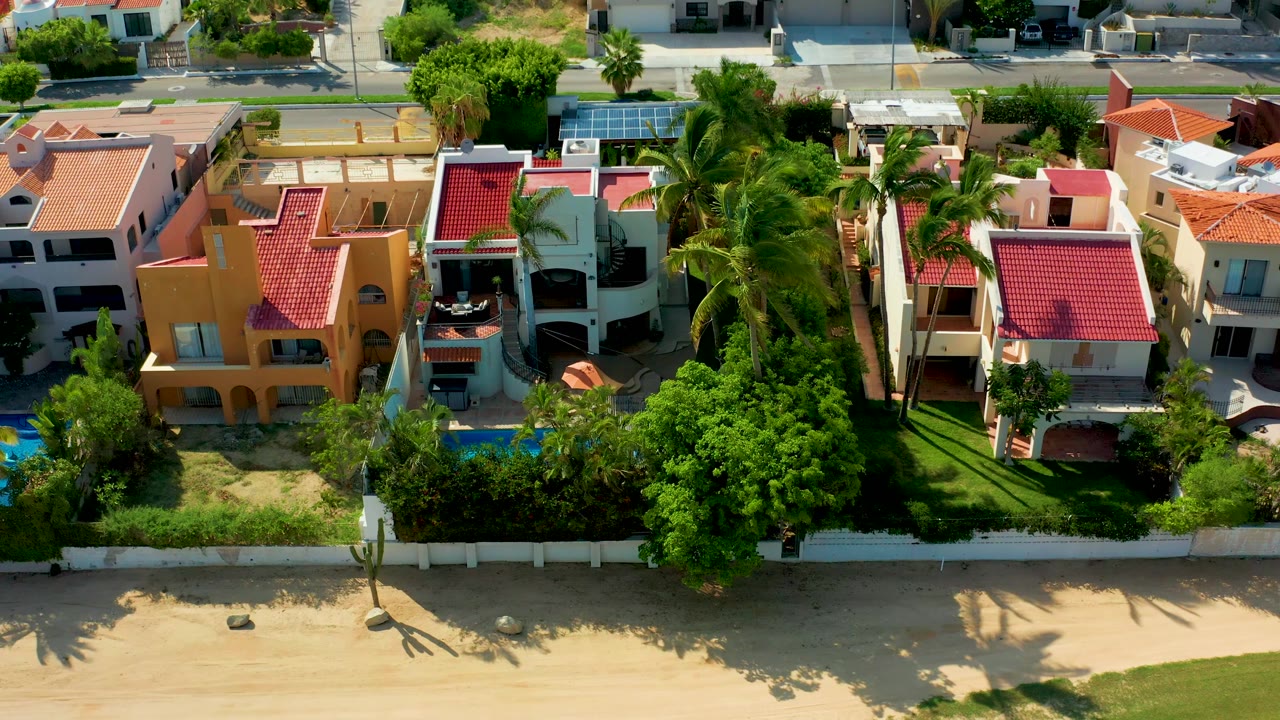 Overhead beachfront residences with palms, red roofs, pools, a white fence, and sand.
