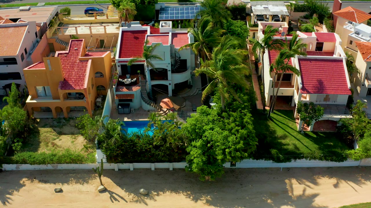 Aerial frame over San Jose Del Cabo beachfront residences with red tile roofs, a small blue pool, palm trees, sandy beach foreground, a street beyond, and no visible people.