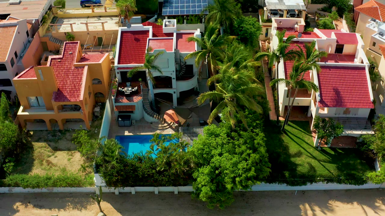 Overhead view of San Jose Del Cabo homes with red tile roofs, a blue pool at lower left, palms and trees, a road at the top, and no visible people.