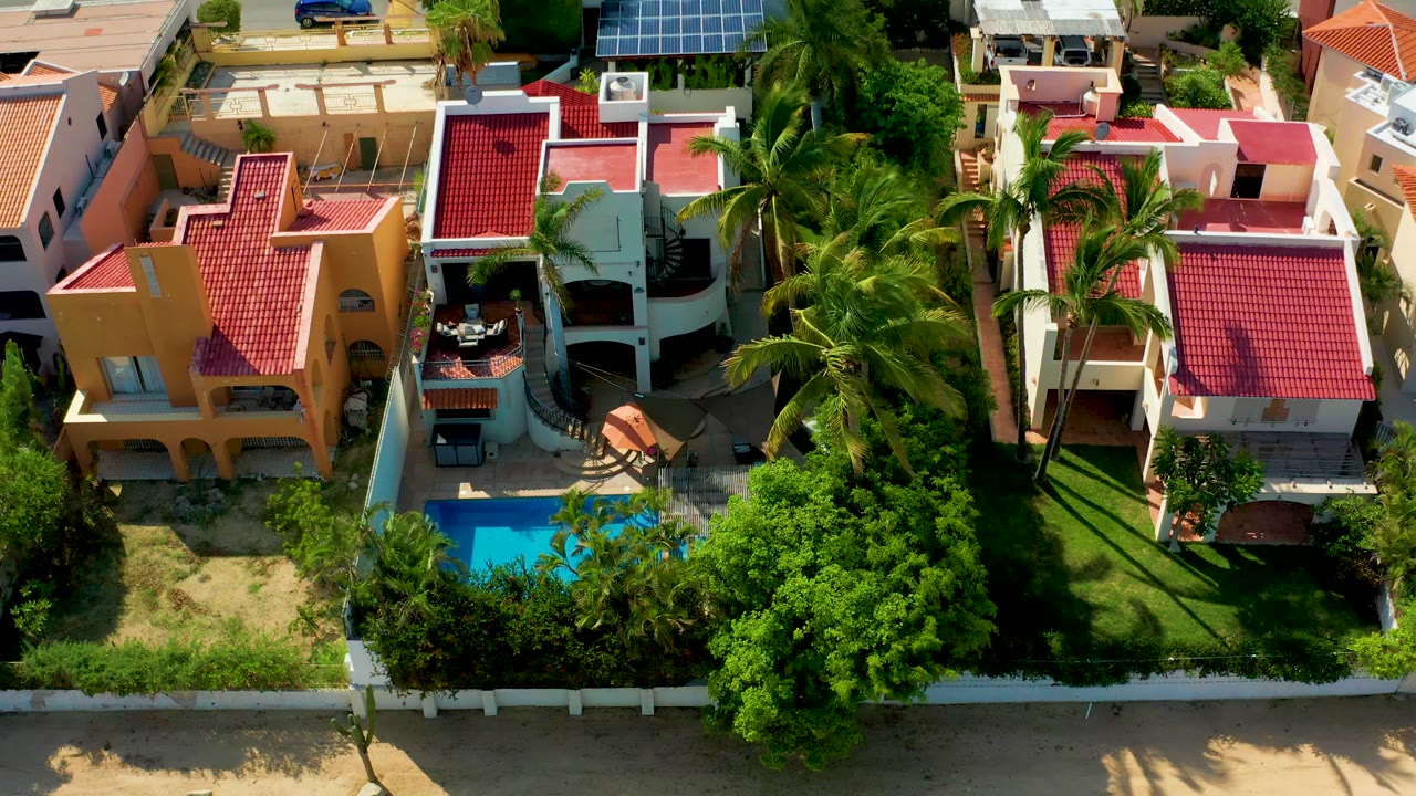 Aerial frame in San Jose Del Cabo showing the same residential block slightly farther along, with palm fronds over the yards, a blue pool near the lower edge, and no visible people.