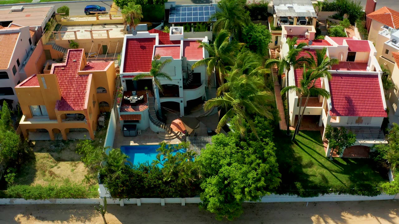 High overhead San Jose Del Cabo residential view with a red-roofed home dominating the right side, a pool partly framed by trees below, and no visible people.