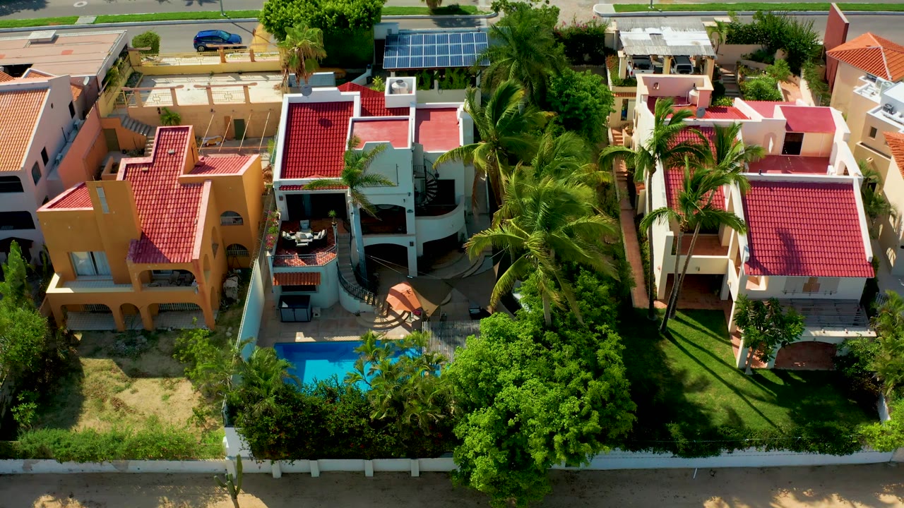 Drone-like overhead frame of houses, pool, palms, road, and fence.