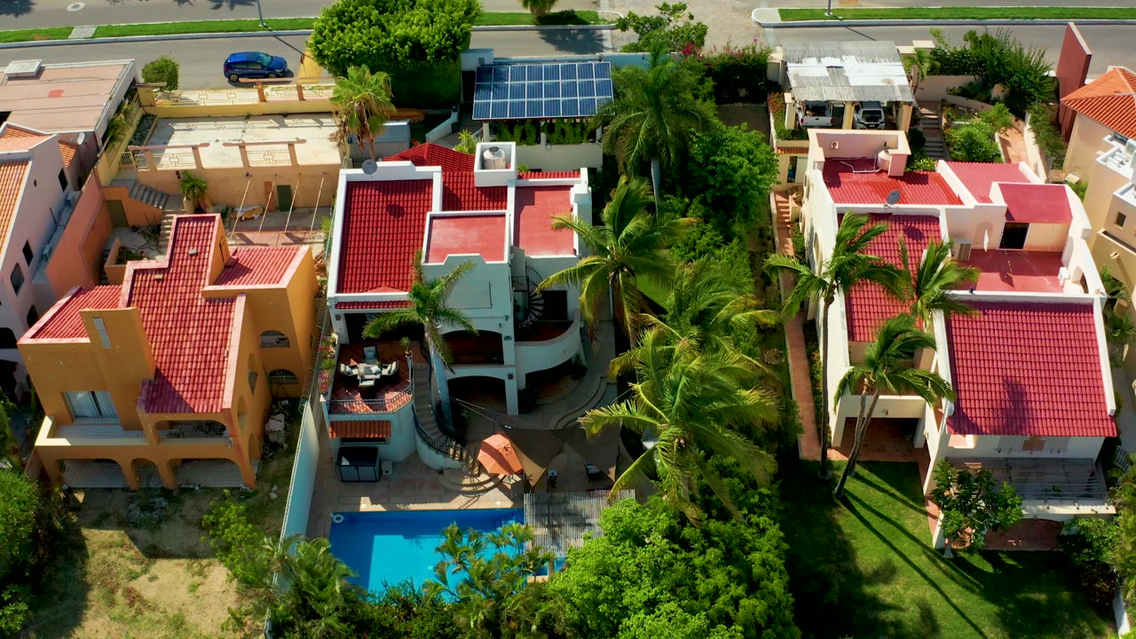 Opening overhead frame in San Jose del Cabo showing red roof homes, a bright blue pool, palms, a street edge, and a blue car with no visible people.