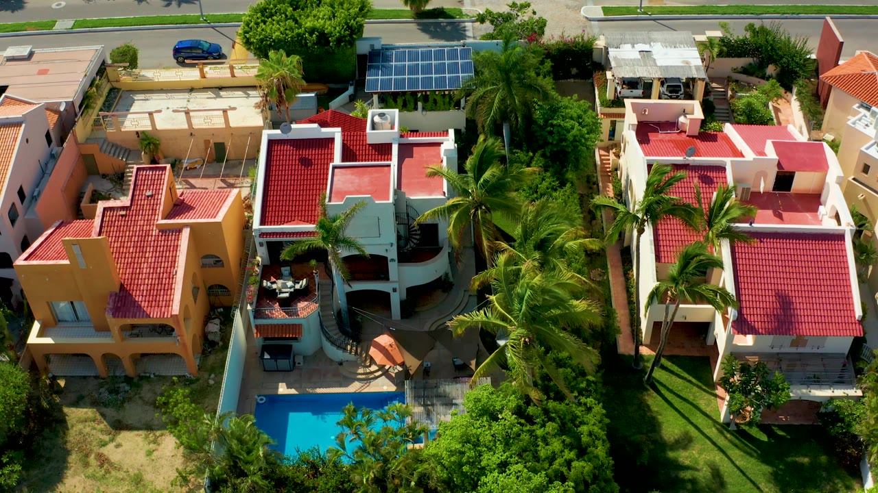 Early aerial frame in San Jose del Cabo with red tile roofs, white walls, a blue swimming pool, palms, solar panels, and no visible people.