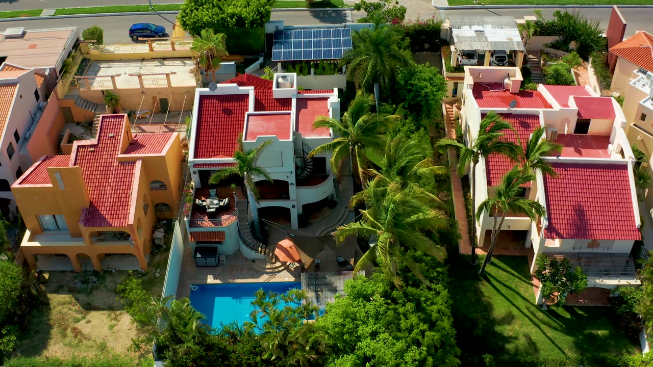 Aerial frame in San Jose del Cabo with the blue car still near the street, red roof homes below it, dense palms to the right, and no visible people.