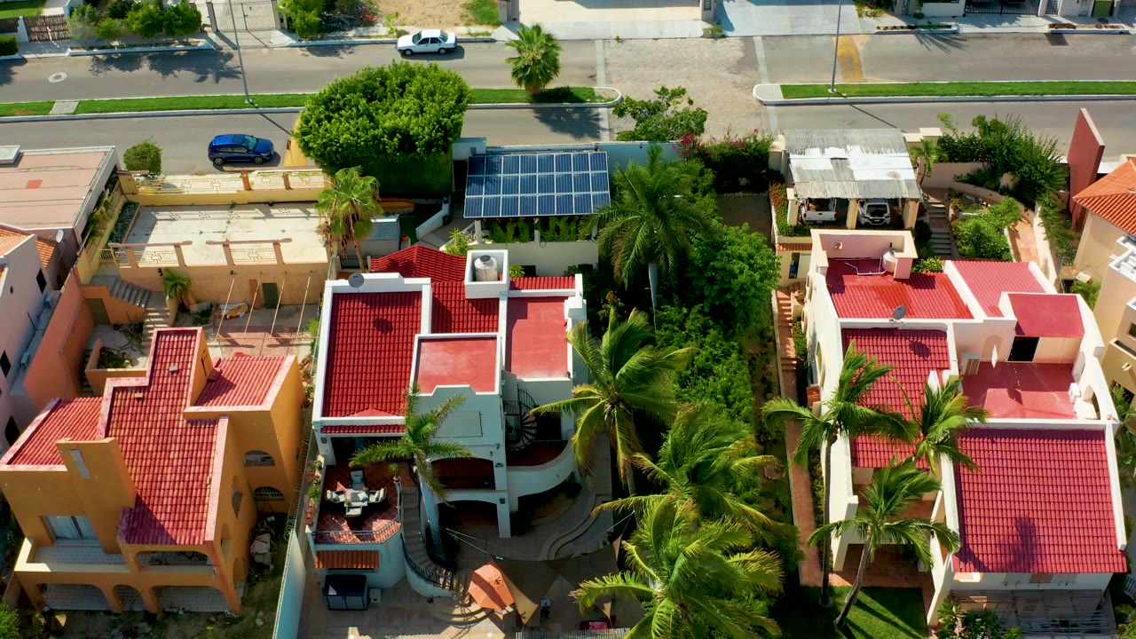 San Jose del Cabo residential roofs fill the frame, with a parked blue car on the upper street, a white car on the road, palms between homes, and no visible people.