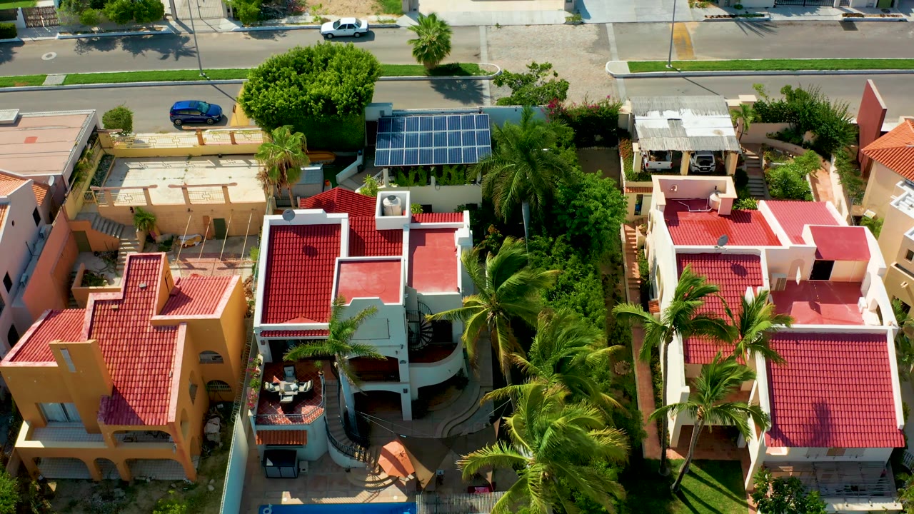 A steep aerial view shows neighboring San Jose del Cabo houses, red roofs, dense palms, a blue parked car near the top road, a white car farther along the street, and no visible people.