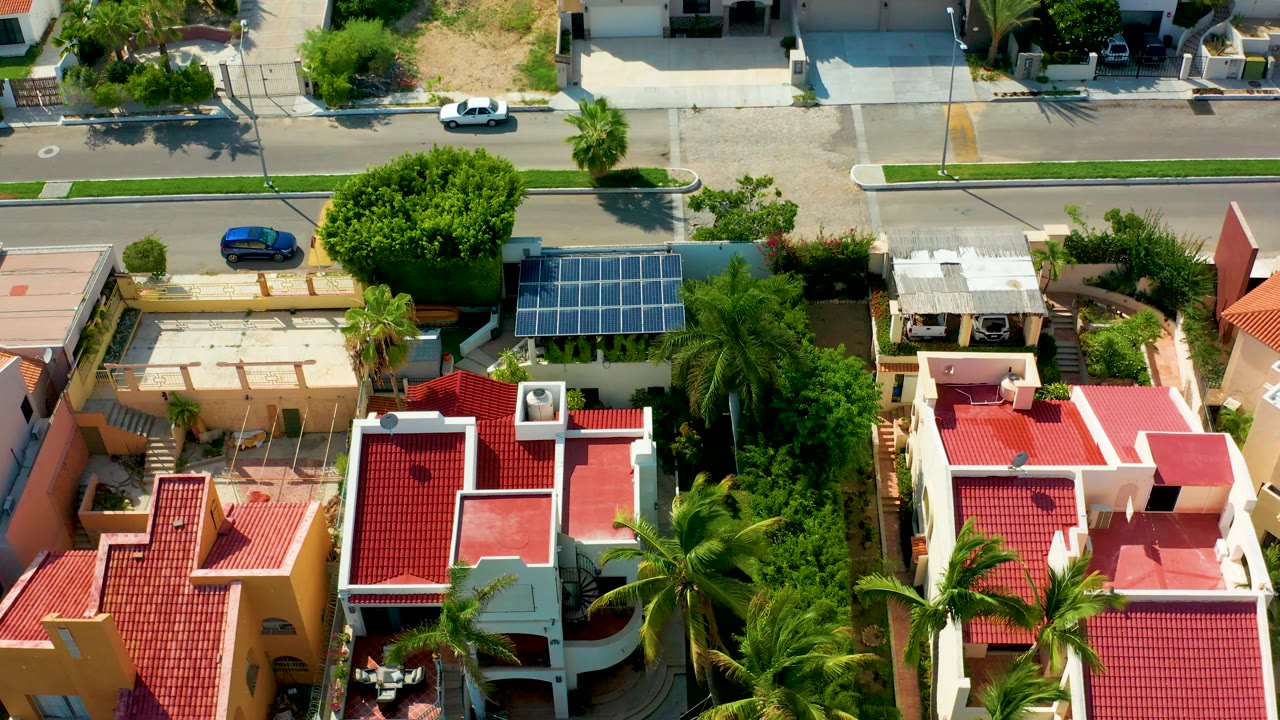 Aerial frame over San Jose Del Cabo homes with red tile roofs, palm trees, a rooftop solar panel array near the upper center, a street at the top, and no visible people.