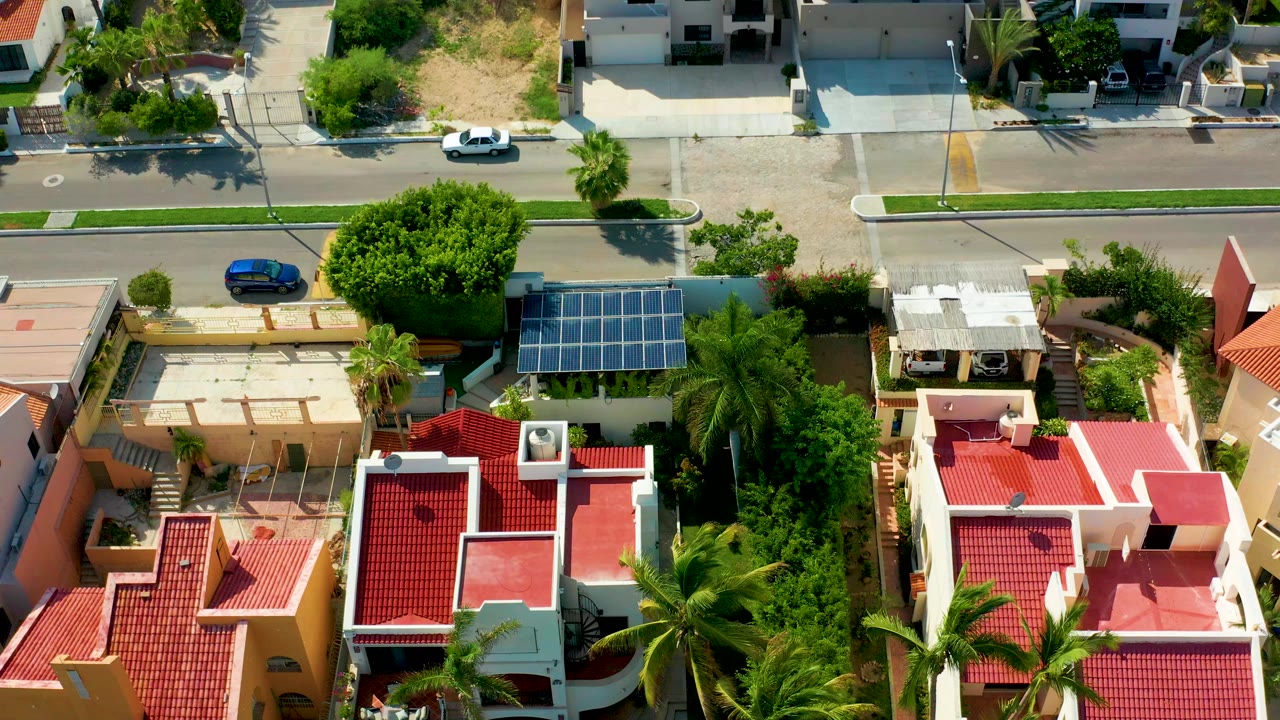 Overhead view in San Jose Del Cabo showing red-roofed homes, palm trees, a top street, solar panels, and no visible people at the start of the clip.
