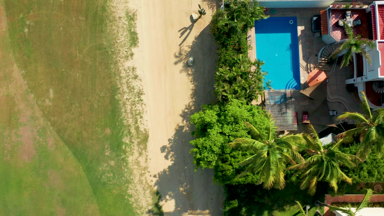 Top-down aerial frame in San Jose del Cabo showing a blue pool, palm trees, garden planting, pale dirt path, green field, and two small visible people near the upper path.