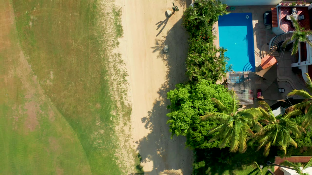 Overhead San Jose del Cabo frame showing two small people and their shadows near the path, with the blue pool, palms, garden shrubs, and green field arranged around them.