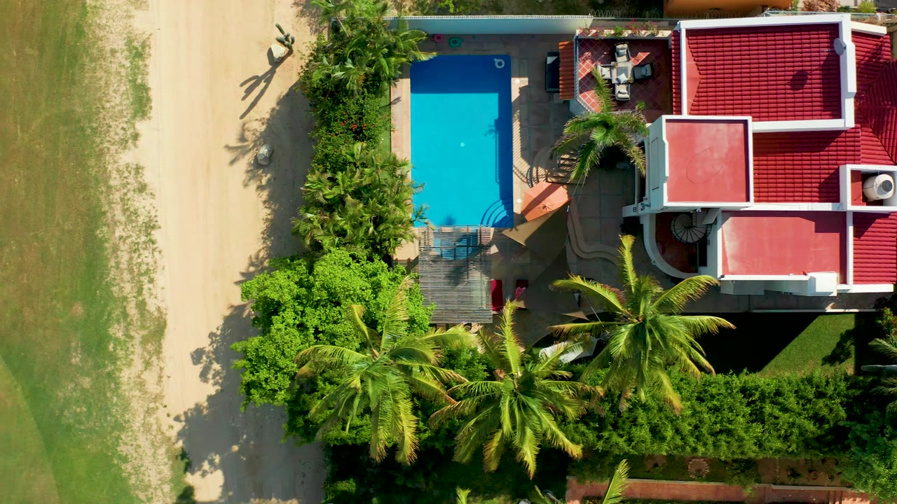 At 0:00, a top-down aerial frame in San Jose del Cabo shows a red-roofed house beside a bright blue pool, with palms, dense greenery, a sandy strip, and grass around it.