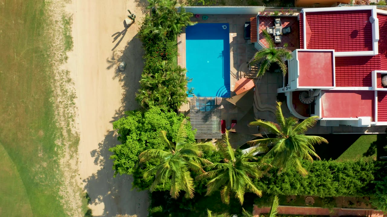 At 0:02, the overhead San Jose del Cabo frame shows the pool along the house edge, red roof sections to the right, palm fronds below, sand to the left, and a small person near the pool area.