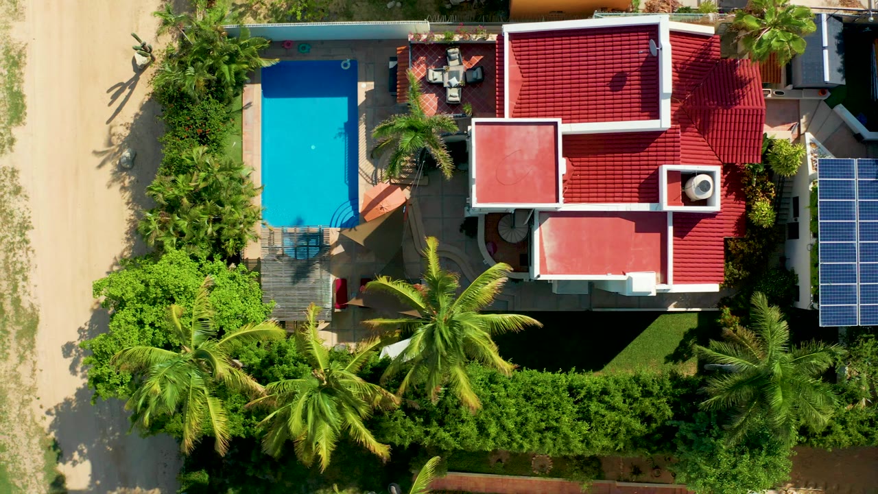Overhead frame in San Jose del Cabo showing a red-roofed house beside a bright blue pool, palm trees, sandy ground, garden hedges, patio furniture, and a solar panel at the right edge, with no visible people.
