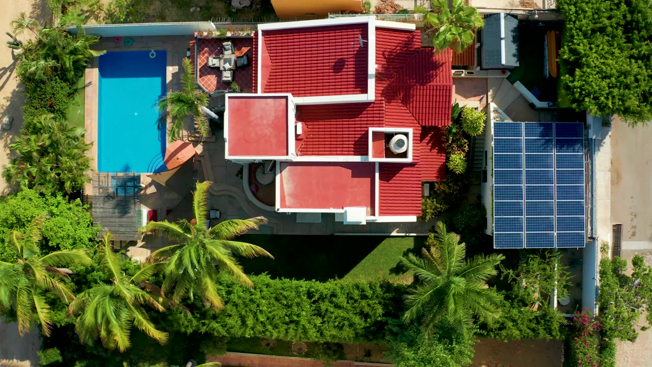 Straight-down aerial frame in San Jose del Cabo showing a red-roofed residence, blue pool on the left, solar panels on the right, palms, hedges, and tiny people near the patio area.