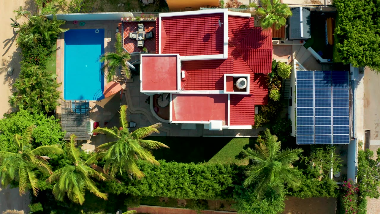 Top-down frame over San Jose del Cabo with a red-roofed residence, bright pool, blue solar panel grid, palm trees, trimmed hedges, paved paths, and tiny people visible near the outdoor seating.