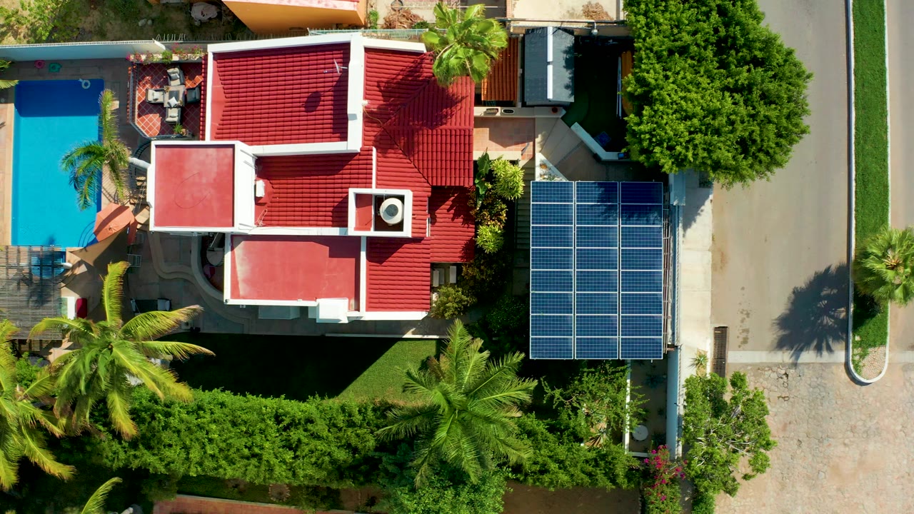 Vertical aerial frame in San Jose Del Cabo showing a red-roofed residential property with a turquoise pool on the left, solar panels beside the driveway on the right, palms and hedges around the yard, and no visible people.