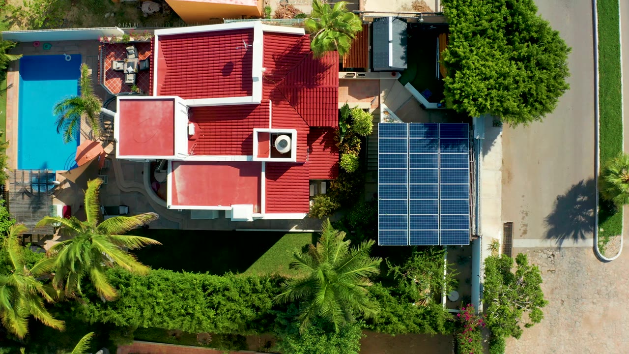 Overhead frame in San Jose Del Cabo with the pool and patio visible at left, the red-roofed house across the center, solar panels along the driveway at right, surrounding palms, and no visible people.