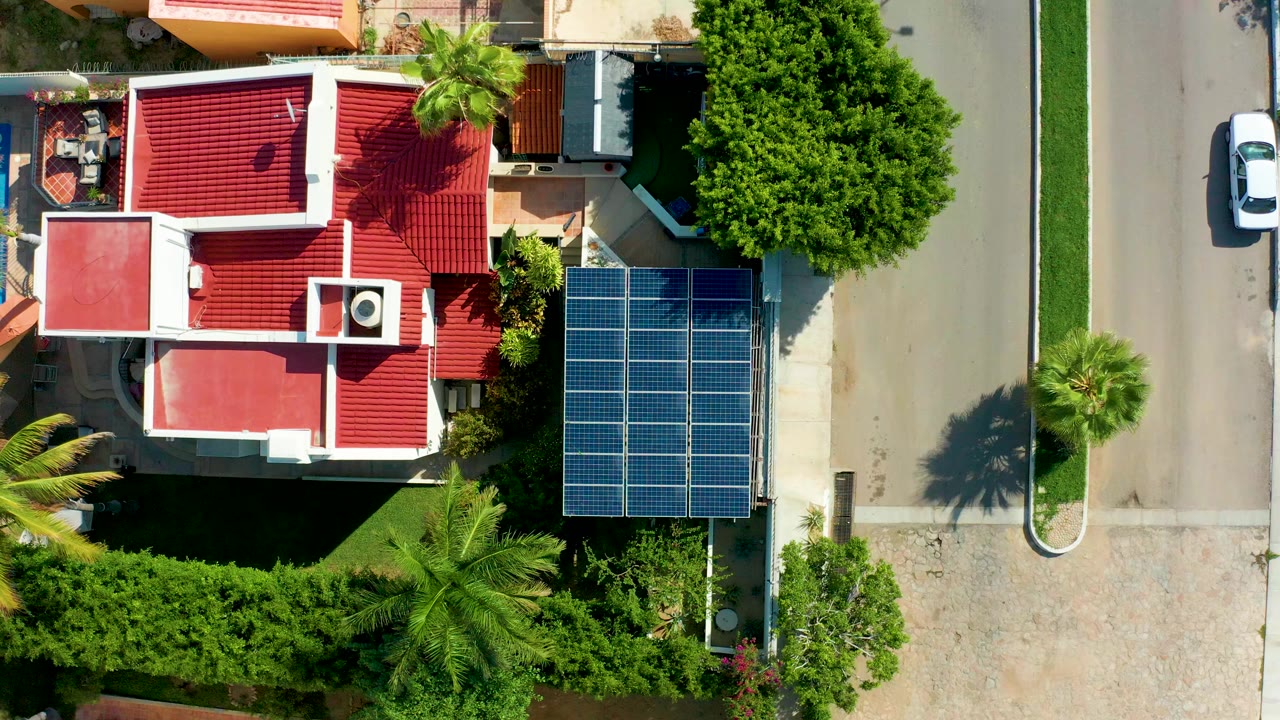 Top-down aerial frame in San Jose Del Cabo, Mexico showing a red-roofed house beside blue solar panels, green trees, a pale street, a parked or slow white car near the road edge, and no visible people.