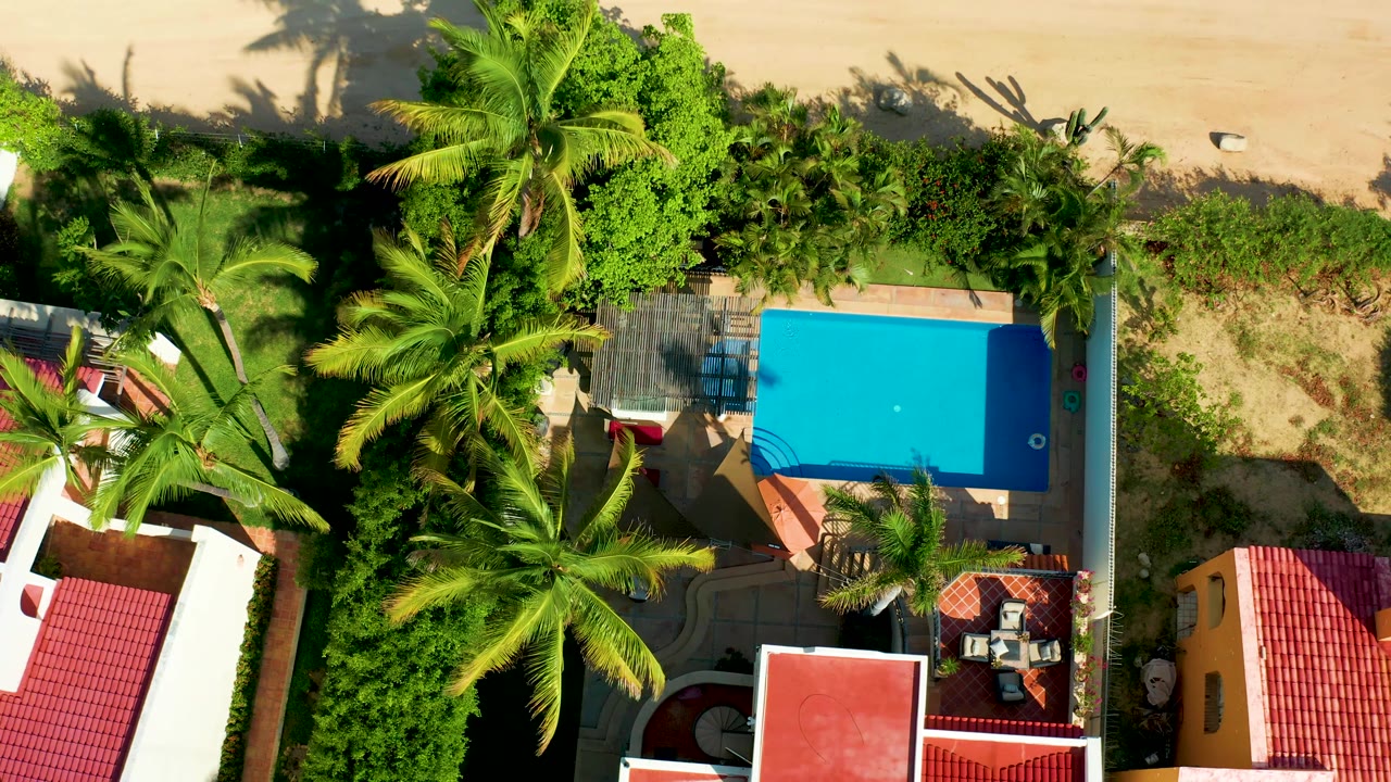 San Jose Del Cabo aerial frame showing the pool and deck framed by palms, red roofs and white walls below, a band of sandy beach above, and no visible people.