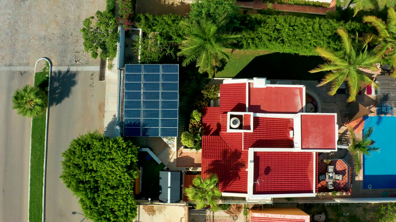 Aerial view of a solar-panel house beside a street, with palms and a blue pool.