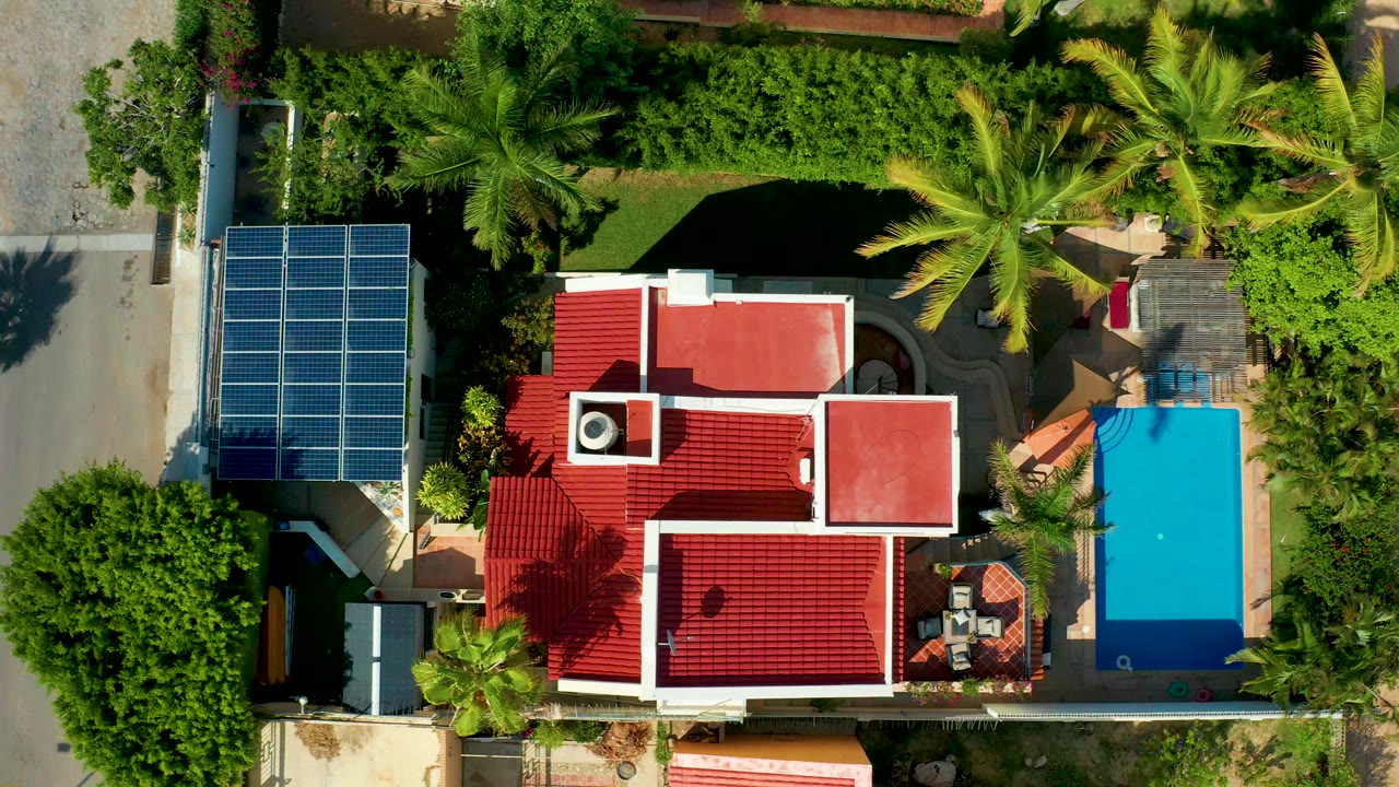 Overhead aerial frame in San Jose del Cabo showing a red roof house, solar panel array, blue pool, street edge, palms, dense greenery, and no visible people.