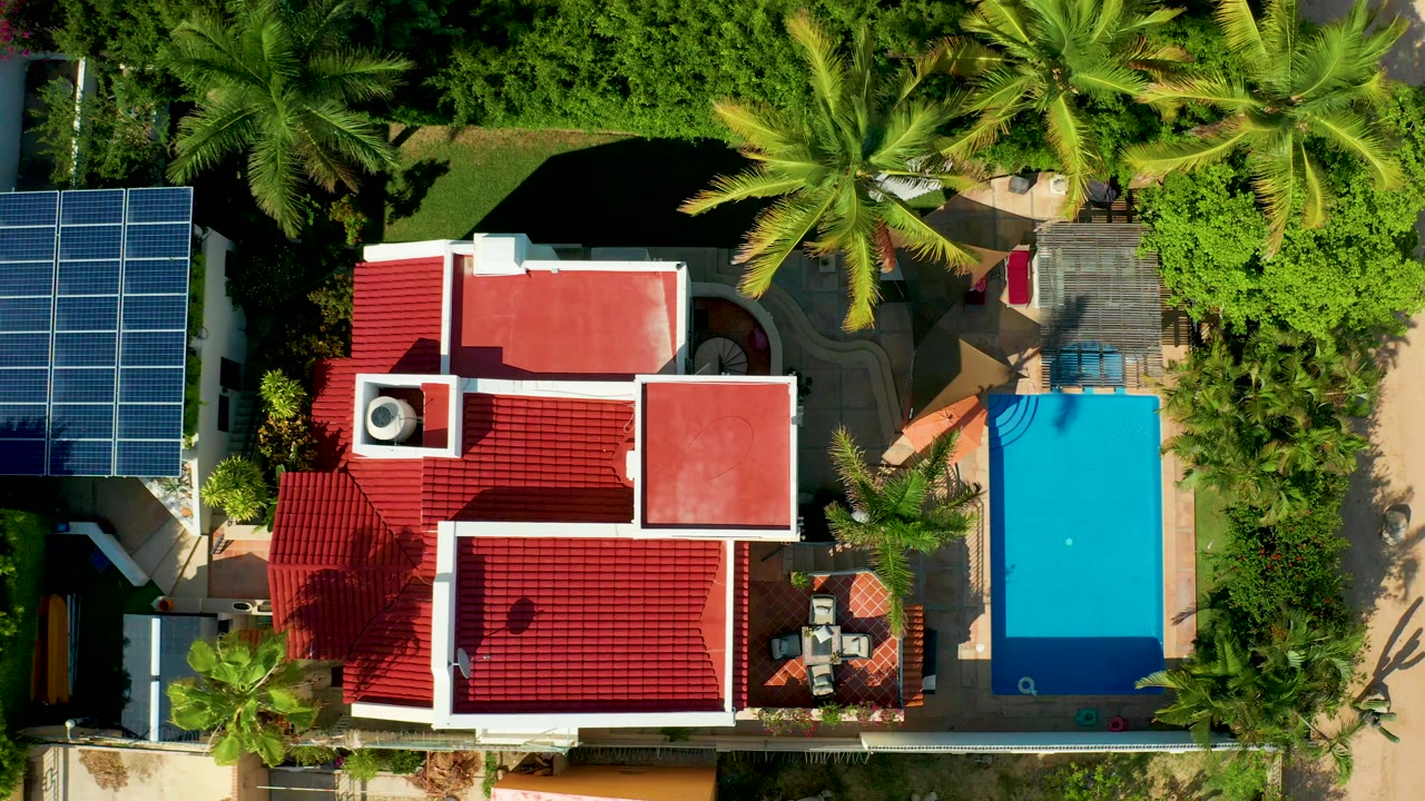 Overhead frame from San Jose Del Cabo showing a red-roofed house centered between dark solar panels and a bright blue pool, with palms, garden paths, hard shadows, and no visible people.