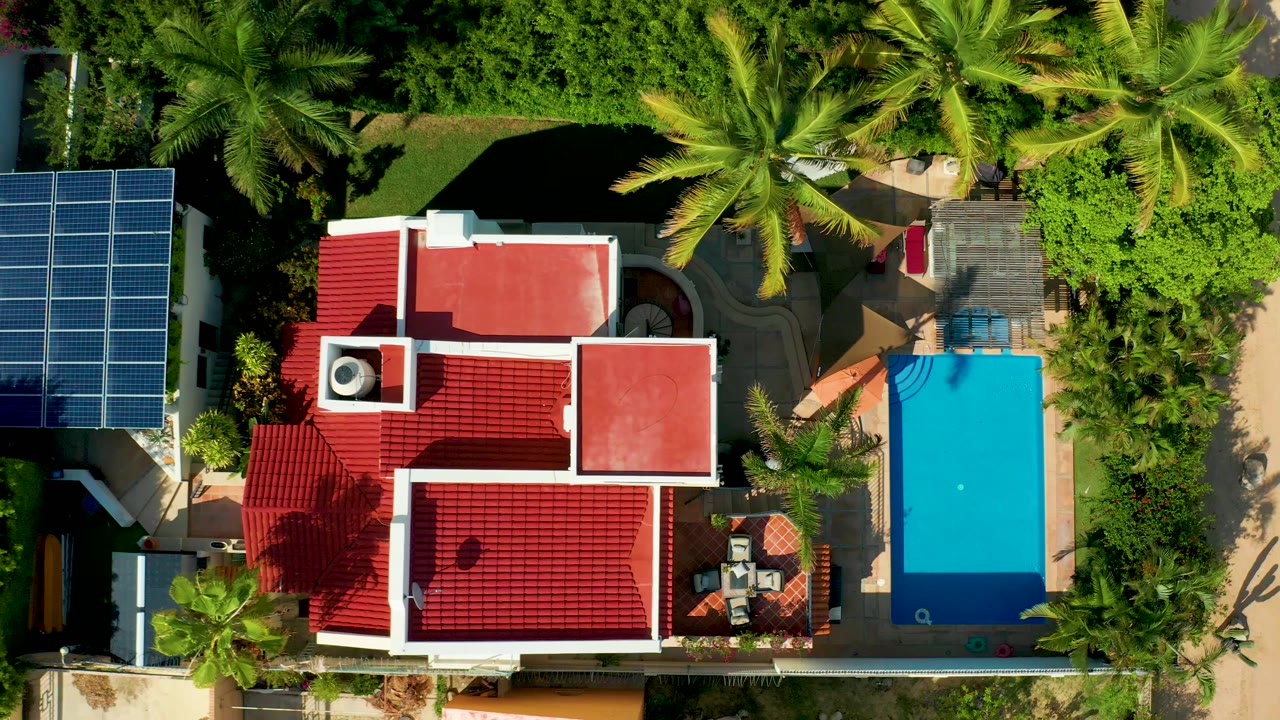 A slightly shifted overhead view in San Jose Del Cabo keeps the red-roofed residence central, with the solar-panel array at left, the rectangular pool at right, palms above, and no visible people.