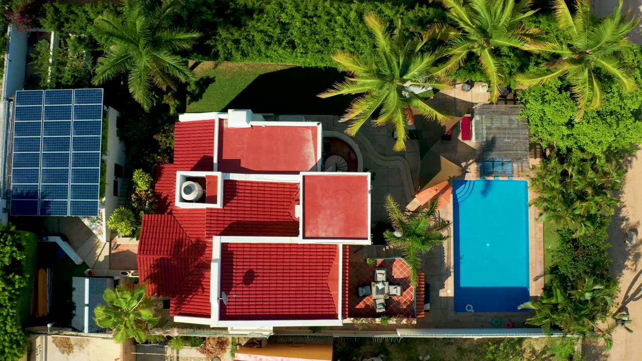 The frame shows the San Jose Del Cabo home from above with more of the right-side planting visible around the pool, while the red roof, solar panels, palm shadows, and no visible people remain clear.
