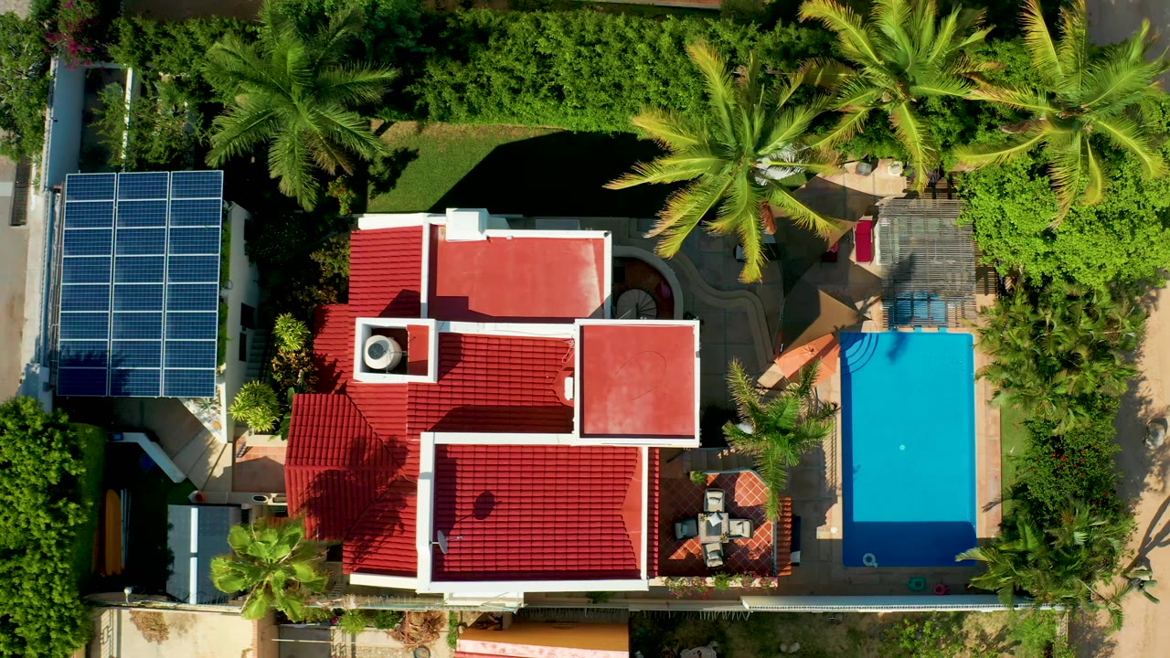 Overhead property view with red roof, bright pool, solar panels, palms, and a small patio area.