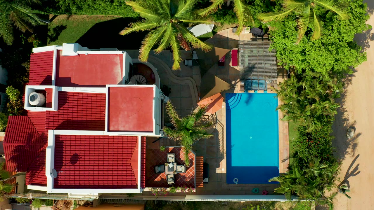 Top-down aerial frame in San Jose del Cabo showing a red-roofed house beside a bright blue pool, palm trees, patio furniture, paved paths, and no visible people.