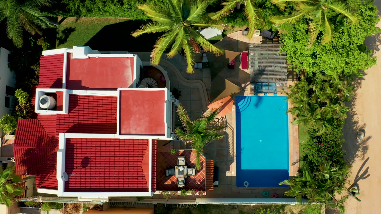 Top-down San Jose del Cabo frame showing the red-roofed residence, rectangular turquoise pool, surrounding palms, garden paths, patio furniture, and no visible people.