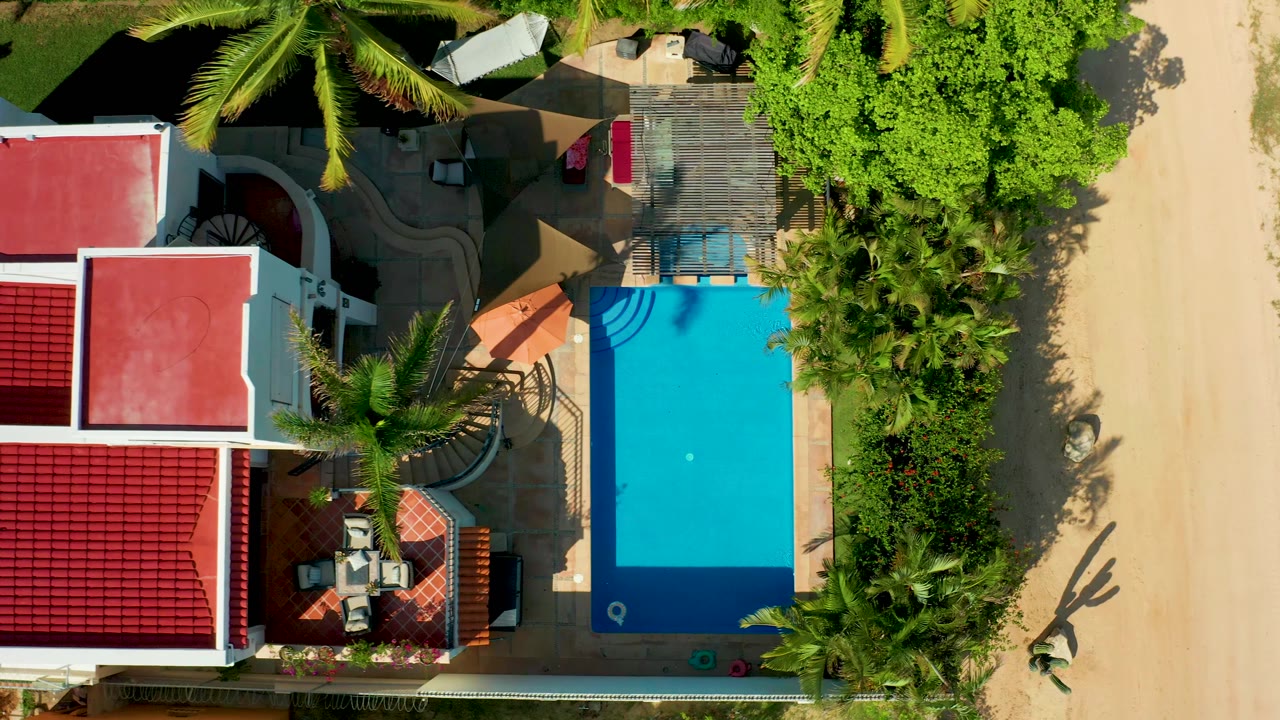 Second aerial frame in San Jose Del Cabo with the pool slightly right of center, red roofs at left, palm shadows across the patio, and no visible people.