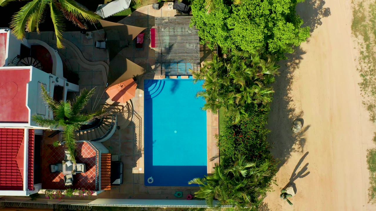 Top-down frame in San Jose del Cabo showing a blue rectangular pool beside red-roofed white buildings, palms, sandy ground, and no visible people.