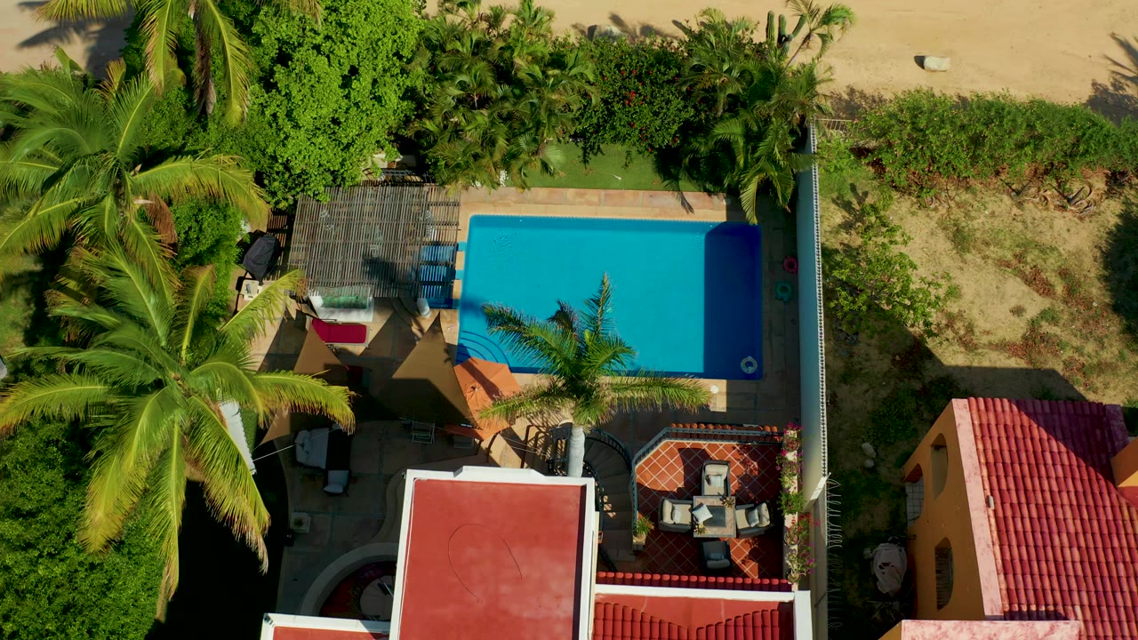 Overhead frame in San Jose del Cabo shows a blue rectangular pool beside palm trees, red-tiled roofs, patio furniture, sandy ground, and no visible people at the start of the clip.
