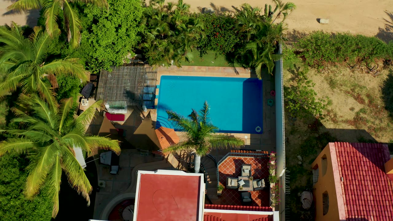 At 0:02, the San Jose del Cabo overhead frame shows the pool centered to the right, palms spanning the left side, roof corners below, sandy ground above, and no visible people.