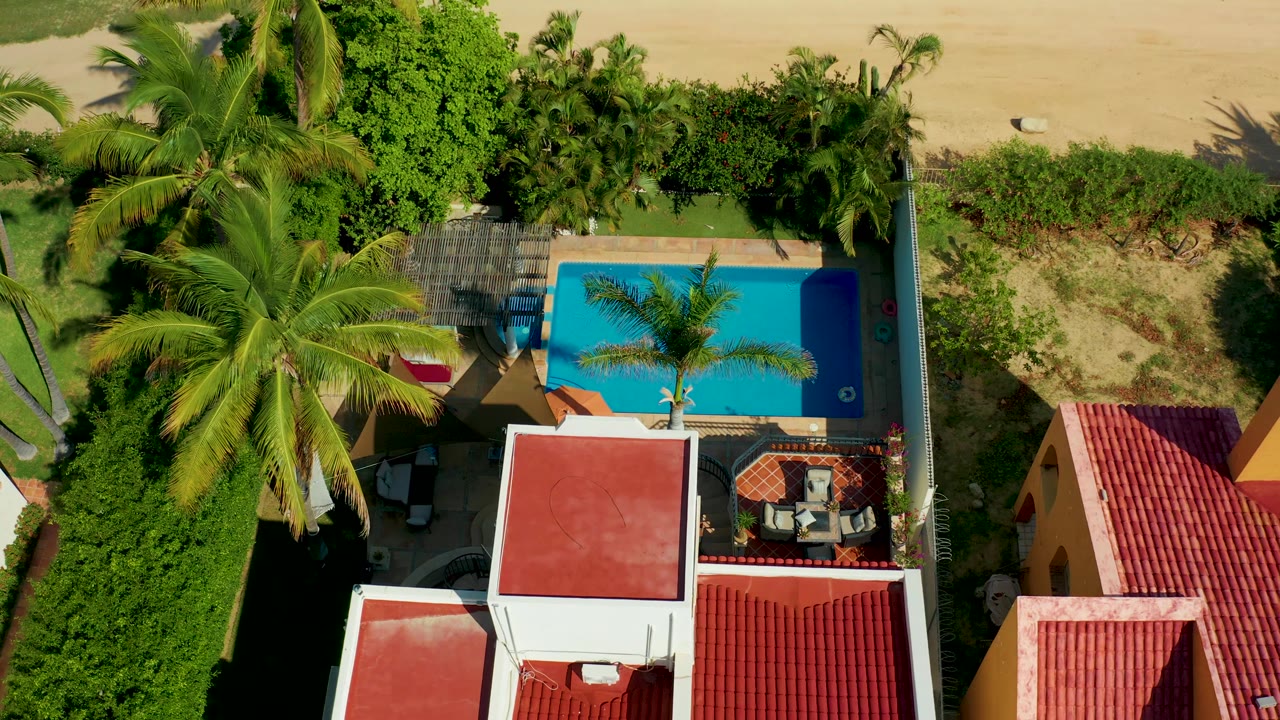 At 0:01, the San Jose Del Cabo overhead view keeps the turquoise pool beside palm shade and red roofs, with beach sand along the upper edge and no visible people.